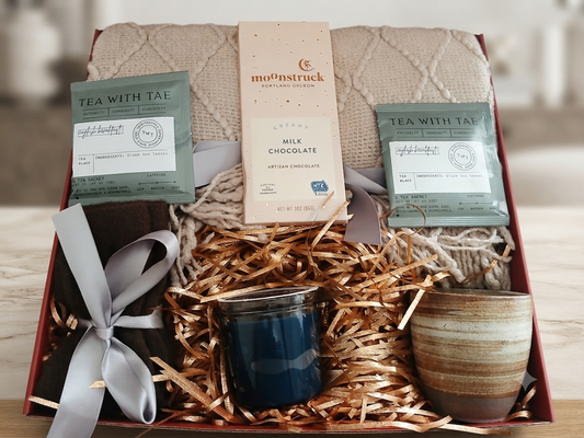 A curated gift box set on a light wood surface, featuring a quilted beige blanket, shredded kraft paper, a dark blue candle, a striped ceramic mug, a Mo'ns Truck milk chocolate bar, and two packages of Tea With Tae tea.