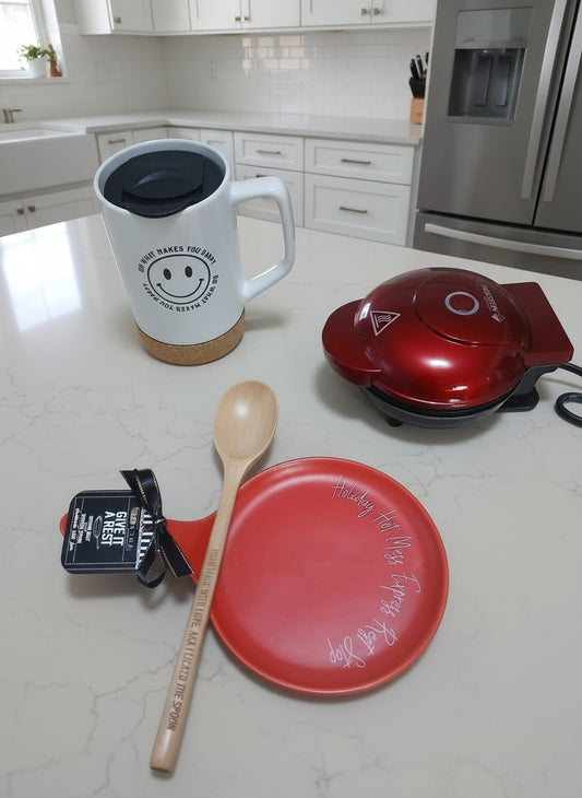 Kitchen items including a mug, wooden spoon, red plate, and waffle maker on a countertop.