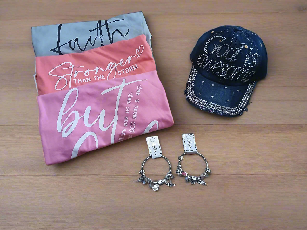 Three t-shirts with motivational phrases, a cap with 'God is awesome' text, and two bracelets on a wooden surface.