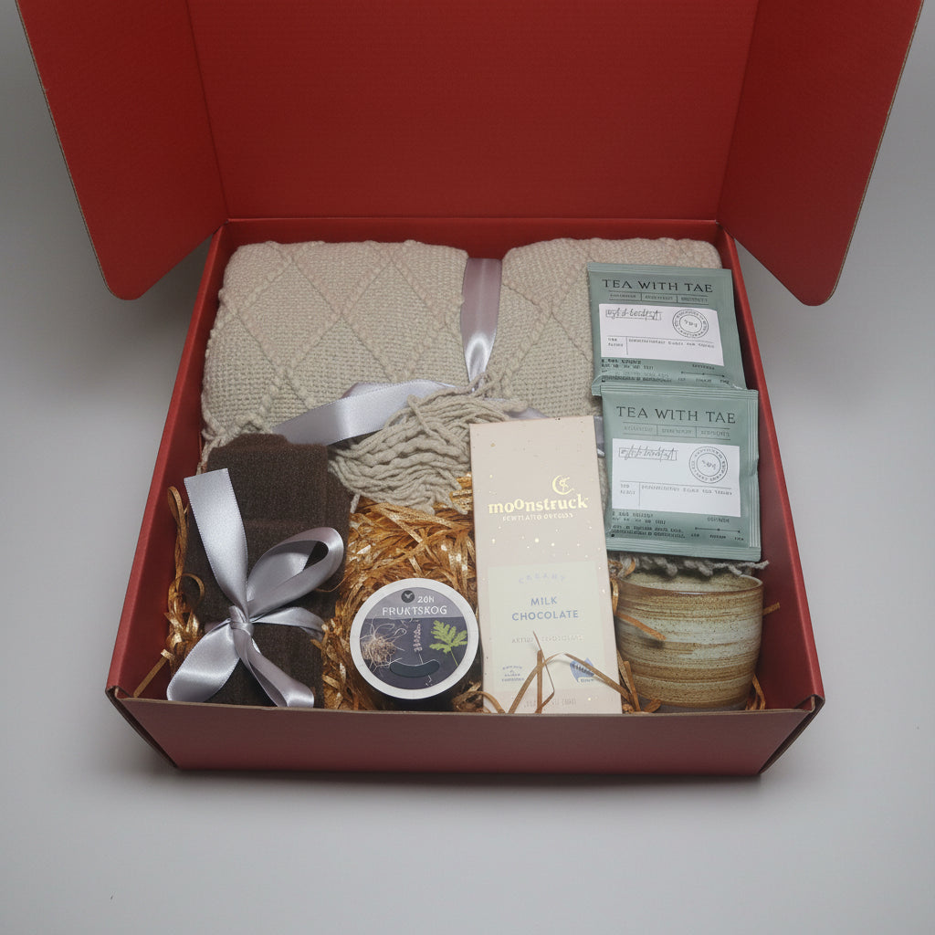 An open red gift box containing a beige knitted blanket, tea bags, a small round tin, a wrapped chocolate bar, and a jar, arranged in gold shredded paper on a light gray background.