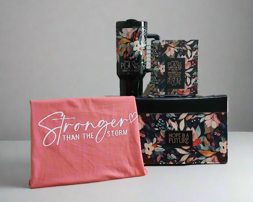 Floral-patterned books on a wooden surface with a pink towel reading 'Stronger Than The Storm' in the foreground.