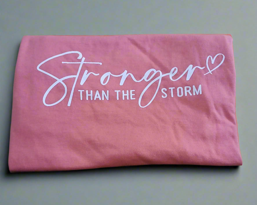Pink t-shirt with 'Stronger Than The Storm' text on a wooden table