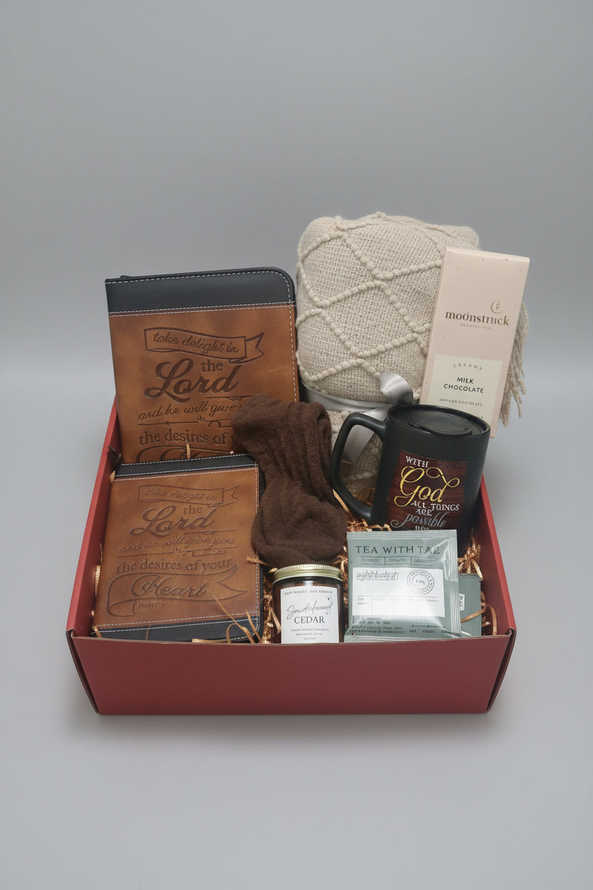 A curated gift box set on a wooden table containing a brown leather wallet with 'God is my delight' engraving, a dark green ceramic mug with 'With God all things are possible' text, a brown knit beanie, a candle, a jar of cedar, and tea bags.